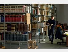 Interno di un archivio o biblioteca con alte scaffalature metalliche piene di volumi rilegati, fascicoli e scatole etichettate. Sul lato destro si vede una postazione di lavoro con sedia girevole e scrivania, in un ambiente illuminato da luce naturale.