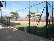 Campo sportivo all’aperto con erba sintetica, recinzione alta in rete metallica e fari per l’illuminazione. Accanto c’è un vialetto pavimentato e sullo sfondo si vedono palazzi residenziali e alcuni alberi.
