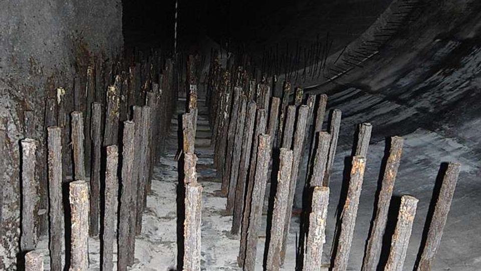 Interno di un grande tunnel o condotta scura, con pavimento in calcestruzzo e file fitte di pali verticali arrugginiti disposti in corridoi. In primo piano si vede una barriera a nastro bianco e rosso che delimita l’area.