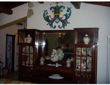 Interno rustico con soffitto a travi in legno e parete bianca decorata da un grande motivo floreale dipinto. Due vetrine in legno scuro con ante in vetro espongono ceramiche e piatti decorati, con un mobile centrale e specchio sullo sfondo.
