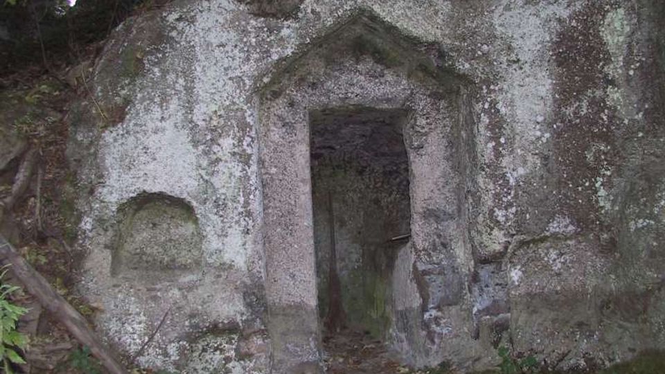 Ingresso rettangolare scavato nella roccia, con architrave a forma di timpano e pareti in pietra grezza. La facciata è coperta da licheni e muschio, inserita in un’area boscosa con vegetazione ai margini.
