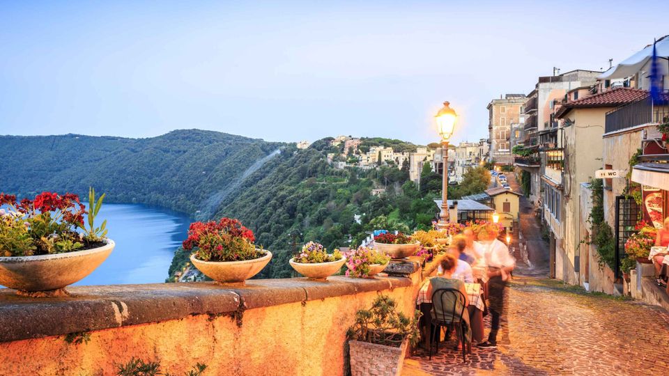 Terrazza panoramica su una strada acciottolata con muretto in pietra e vasi di fiori, affacciata su un lago circondato da colline boscose. Sul lato destro si vedono edifici del borgo arroccato e un lampione acceso lungo la via.