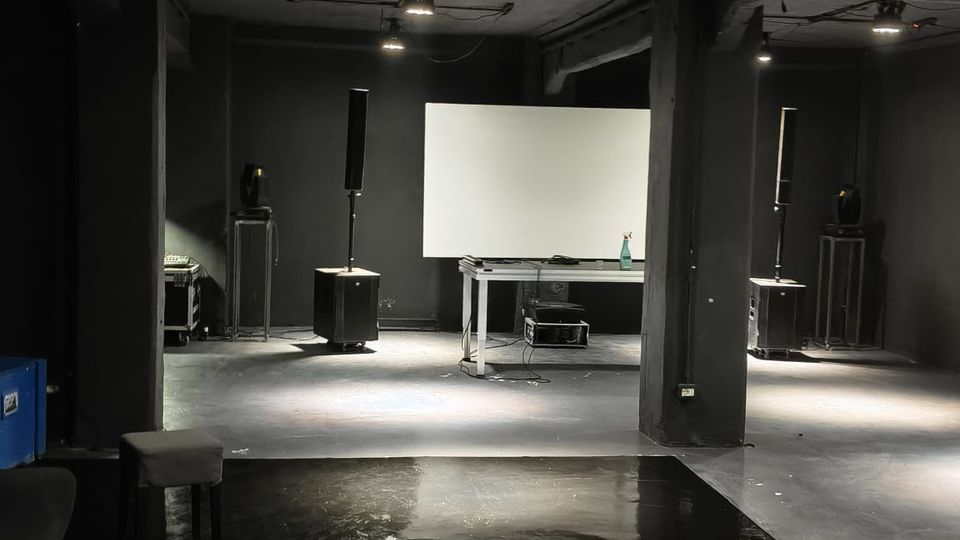 Interno di uno spazio industriale scuro con soffitto in cemento a vista e lampade sospese, pareti nere e pavimento lucido. Al centro ci sono un grande schermo bianco su un tavolo e diverse casse acustiche su supporti distribuite nella sala.