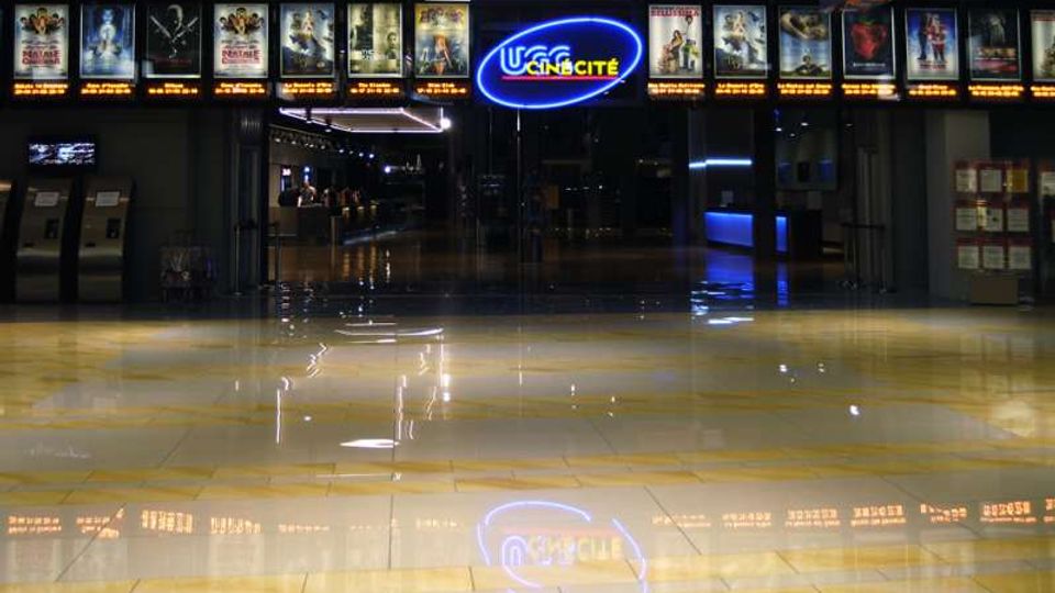 Ingresso di un cinema con pavimento lucido in piastrelle chiare e riflessi, corridoio centrale illuminato e insegna al neon “UCI CINECITTÀ” sopra l’entrata. Lungo la parete superiore sono visibili vetrine illuminate con locandine di film e, ai lati, chioschi automatici e bacheche informative.