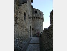 Vicolo stretto in pietra con pavimentazione a ciottoli e muri grezzi, illuminato da una lanterna a parete. In fondo si vede una grande torre cilindrica in muratura con beccatelli e piccole aperture, con un vaso di fiori vicino al passaggio.