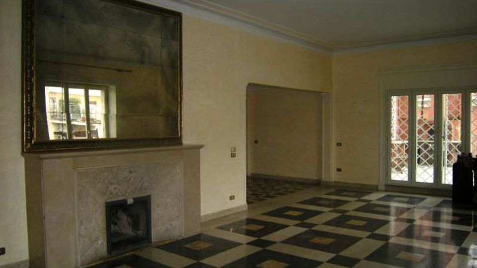 Interno di un ampio soggiorno con pavimento a scacchiera in marmo nero e chiaro, camino in pietra con grande specchio sopra e pareti color crema. Sul lato destro ci sono porte finestre con grate decorative che danno su un balcone, e un’apertura conduce a un’altra stanza con lo stesso pavimento.