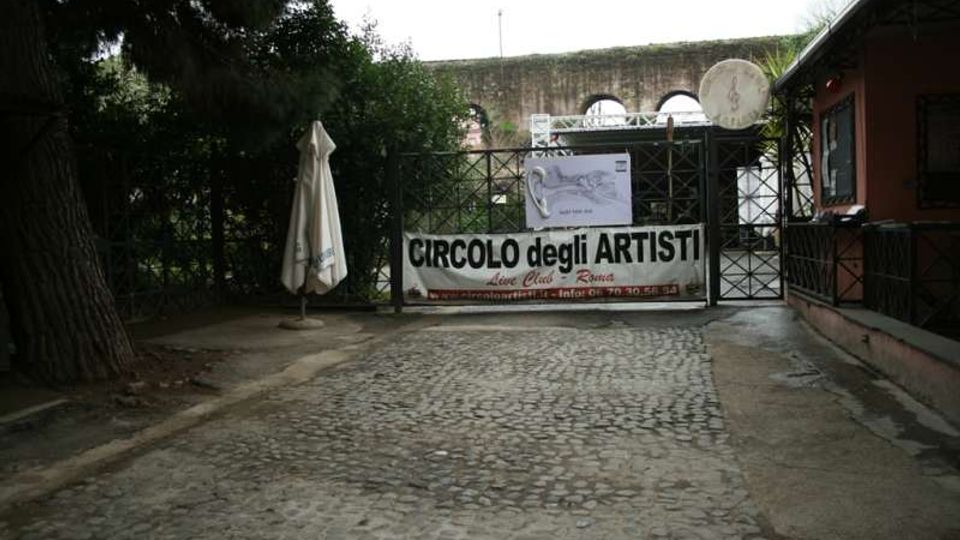 Ingresso con cancello in ferro e strada in ciottoli, con un grande striscione “Circolo degli Artisti” appeso davanti. Ai lati ci sono alberi e un ombrellone chiuso, e sullo sfondo si intravede un muro in pietra con arcate.