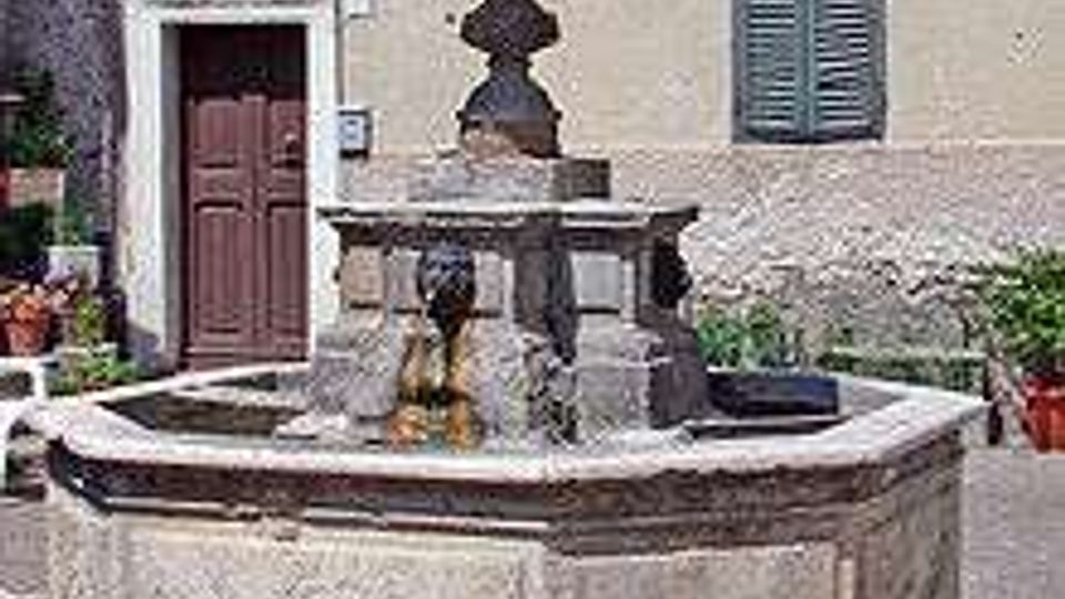 Fontana in pietra a più vasche con colonna centrale, situata in una piccola piazza lastricata davanti alla facciata di un edificio con porta in legno e persiane verdi.