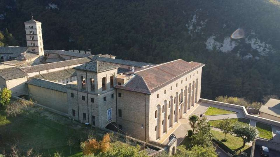 Complesso monastico in pietra su un pendio, con edifici a tetti in coppi e un alto campanile quadrato. Davanti si vedono un cortile e giardini terrazzati, con una collina boscosa sullo sfondo.