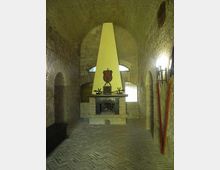 Interno di una sala in mattoni con soffitto a volta e pavimento in laterizio a spina di pesce, con un grande camino centrale sormontato da una cappa triangolare chiara. Ai lati si vedono nicchie ad arco, finestre con vetri opachi e una parete illuminata da una lampada.