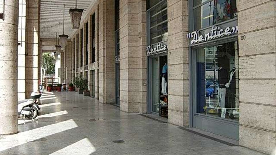 Porticato urbano con alti pilastri in pietra e soffitto a travi, che crea un lungo corridoio ombreggiato. Sul lato destro si vedono vetrine di negozi con ampie finestre, mentre il pavimento in pietra è attraversato da fasci di luce e ombre.