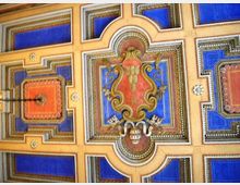 Soffitto decorato con riquadri geometrici blu e arancioni incorniciati da cornici dorate, con medaglione centrale ornato da motivi floreali e araldici. Al centro è visibile un punto luce pendente.