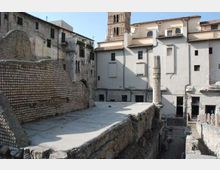 Resti archeologici in pietra con una lunga passerella sopraelevata e un alto muro a mosaico sul lato sinistro. Sullo sfondo si vedono edifici storici con facciate intonacate e un campanile in mattoni che svetta sopra i tetti.