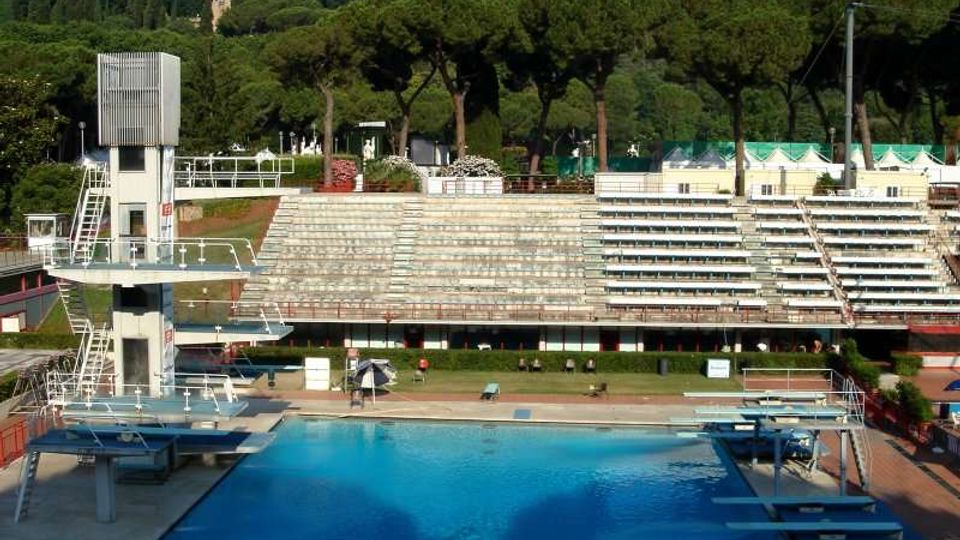 Piscina olimpionica all’aperto con piattaforme e trampolini per tuffi e una torre con scale sul lato sinistro. Sullo sfondo si vedono ampie gradinate in cemento e una fila di pini con una collina boscosa dietro.