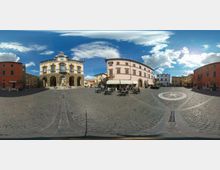 Piazza lastricata in pietra con ampio spazio aperto, circondata da edifici storici a più piani; a sinistra un palazzo con portici ad archi e facciata decorata, al centro un edificio curvo con tende da caffè e tavolini all’aperto. Sul selciato è visibile un motivo circolare e sullo sfondo cielo azzurro con nuvole.