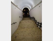Scala in pietra che scende in un corridoio sotterraneo con pareti bianche e soffitto a volta. Corrimano e ringhiere nere lungo i lati conducono a un arco che apre su uno spazio più buio illuminato da piccole luci.