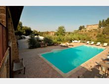 Piscina rettangolare all’aperto con bordo in pietra e pavimentazione in cotto, circondata da lettini e ombrelloni su una terrazza. Sullo sfondo si vede un paesaggio collinare con alberi e un edificio in pietra, accanto a una struttura rustica con balcone.