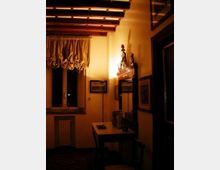 Interno di una stanza in stile tradizionale con soffitto a travi in legno, finestra con tende arricciate e pareti illuminate da luce calda. In primo piano un tavolo con oggetti decorativi e uno specchio incorniciato sopra, con quadri appesi e una porta scura sul lato destro.