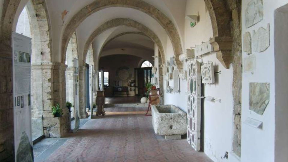 Corridoio interno in stile storico con archi in pietra e soffitto a volte, pavimento in mattoni e pareti bianche. Lungo le pareti sono esposti pannelli informativi e rilievi in pietra, con una grande vasca in pietra sul lato destro.