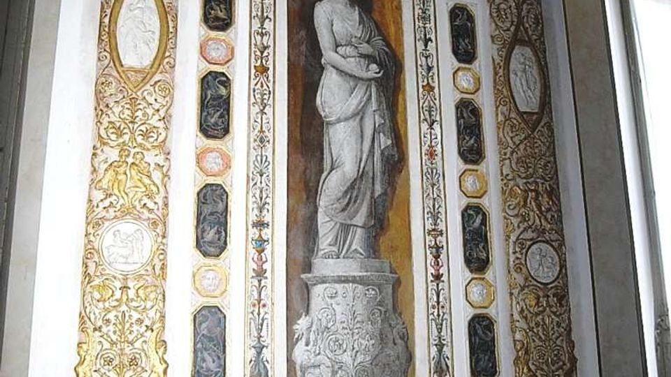 Parete interna decorata in stile classico con pilastri riccamente dorati, archi e medaglioni ornamentali. Al centro, una nicchia con statua dipinta in monocromo incorniciata da fregi, e sotto due pannelli circolari con uccelli, accanto a una finestra laterale.