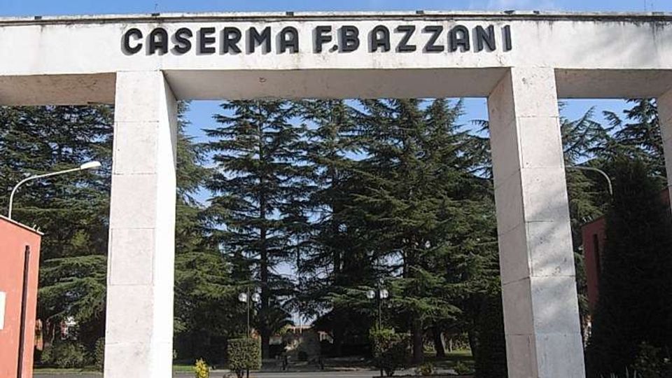 Ingresso di una caserma con grande portale rettangolare in cemento, con la scritta “CASERMA F.B. AZZANI” in alto. Oltre l’arco si vede un viale pavimentato che conduce a un’area interna alberata con alti pini e lampioni.