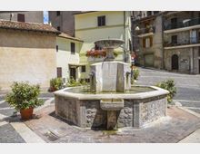 Fontana in pietra al centro di una piccola piazza lastricata, con vasca poligonale e colonna centrale con vaschetta superiore e beccucci da cui scorre l’acqua. Intorno si vedono edifici residenziali a più piani con persiane e balconi, alcuni vasi di piante e una strada in lieve salita sullo sfondo.