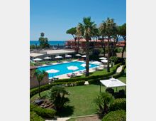 Piscina all’aperto di un hotel con lettini e ombrelloni bianchi disposti lungo il bordo, circondata da prato curato e siepi. Palme e pini marittimi incorniciano l’area, con edifici del complesso e il mare visibile sullo sfondo.