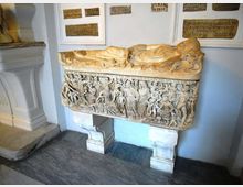 Sarcofago in pietra esposto in una sala museale, con coperchio scolpito a figura distesa e pannello frontale riccamente decorato a bassorilievo con scene e personaggi. Sul muro chiaro dietro sono montate varie lapidi con iscrizioni.
