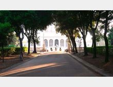 Viale asfaltato in un parco alberato, fiancheggiato da lampioni e siepi, che conduce a un edificio bianco in stile classico con facciata simmetrica e grandi arcate. Sul davanti sono visibili una scalinata e vasi ornamentali lungo la terrazza.