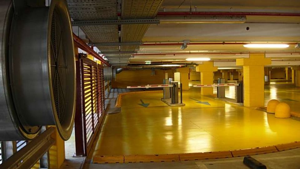 Parcheggio sotterraneo con pavimento giallo lucido e colonne gialle, illuminato da luci al neon e con tubazioni e canaline a soffitto. Si vedono una sbarra di accesso, segnaletica con frecce sulla pavimentazione e una grande ventola industriale sulla sinistra.