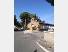 Piccola chiesa in mattoni con facciata semplice, arco sopra l’ingresso e campanile a vela, affiancata da un edificio basso e allungato. L’area circostante è una strada asfaltata con parcheggi e alberi alti (pini) lungo il viale, sotto un cielo sereno.