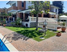 Villa a due piani in mattoni e intonaco bianco con portico e terrazza con balaustre, affacciata su un giardino con prato, albero centrale, lampioncini e vasi. In primo piano si vede un vialetto pavimentato e il bordo di una piscina.