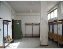 Spogliatoio semplice con pavimento piastrellato e soffitto bianco, dotato di panche in legno, appendiabiti e scaffali lungo le pareti. Una colonna centrale divide lo spazio; sul lato destro ci sono finestre alte che lasciano entrare luce naturale e una porta verde sul fondo.