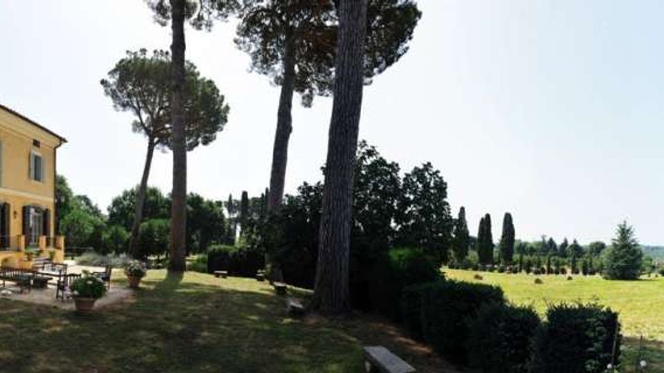 Facciata di una villa gialla a due piani con persiane verdi e terrazza pavimentata con tavoli e vasi. Intorno si estende un ampio giardino con pini alti, siepi e prato che apre sulla campagna con cipressi in lontananza.