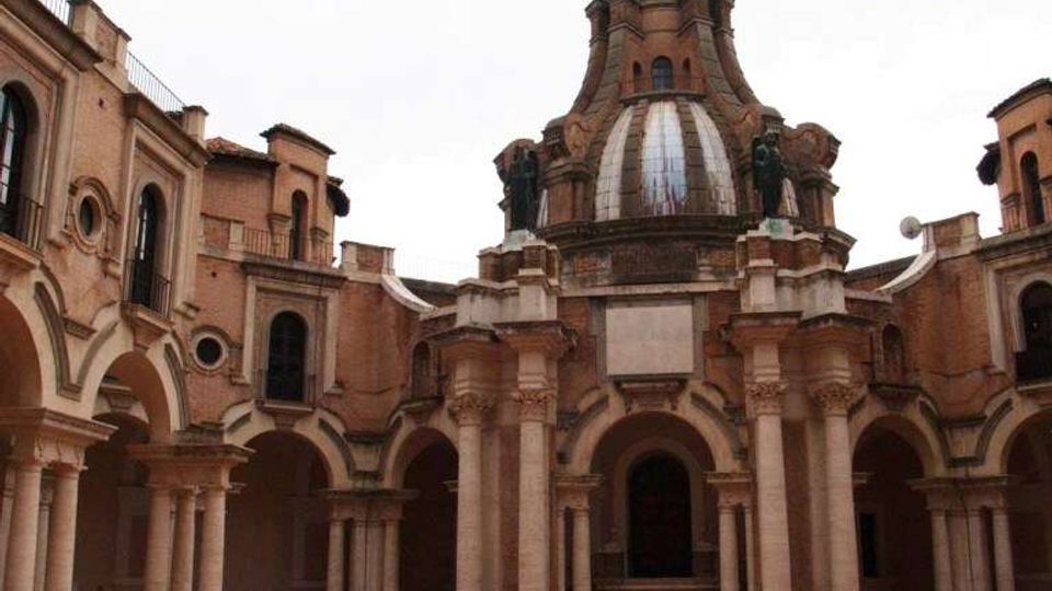 Cortile interno di un complesso storico in mattoni, con portici ad archi e colonne su più lati e una pavimentazione in laterizio a disegni geometrici. Al centro si vede una struttura con cupola e torretta, affiancata da elementi scultorei e aperture ad arco.