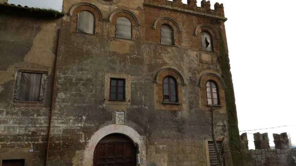 Facciata in pietra di un edificio storico fortificato con merlature, arco d’ingresso e finestre ad arco, con un piccolo orologio sulla parte alta. In primo piano si vede un muro di cinta e un vialetto in pietra sotto un cielo nuvoloso.