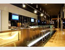 Interno di un bar moderno con lungo bancone in acciaio e fila di sgabelli alti, pavimento lucido e pareti scure con pannelli riflettenti. Luci a sospensione lungo il soffitto e schermi digitali dietro il banco completano l’ambiente.