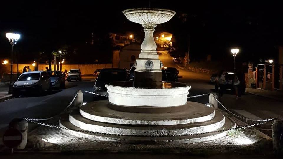 Fontana in pietra illuminata di notte, con vasca circolare su gradoni concentrici e un’alta coppa centrale. Intorno si vede una piccola piazza/rotonda con catene di protezione, auto parcheggiate e lampioni lungo la strada in salita.