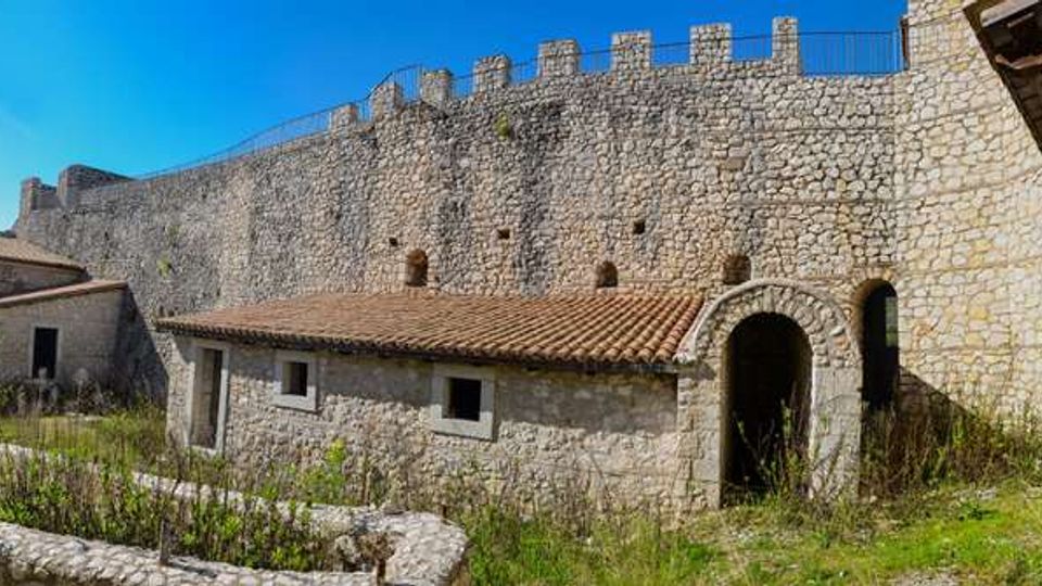 Cortile interno di una fortificazione in pietra con alte mura merlate e una piccola costruzione in pietra con tetto in coppi. Si vedono un arco d’ingresso, edifici annessi e aree erbose con muretti bassi sotto un cielo sereno.
