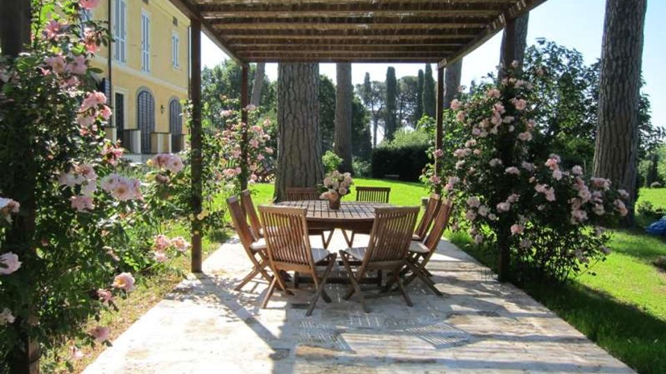 Pergolato in legno su una terrazza in pietra con tavolo rotondo e sedie da esterno, circondato da cespugli di rose. Sullo sfondo si vedono un prato con alti pini e il fianco di una villa gialla con finestre ad arco.