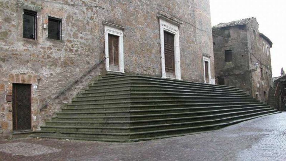 Facciata in pietra di un edificio storico con ampie scalinate in pietra verde che salgono lungo l’angolo fino a un ingresso con cornice chiara. Davanti si apre una piazza pavimentata in mattoni, con altri edifici antichi sullo sfondo.