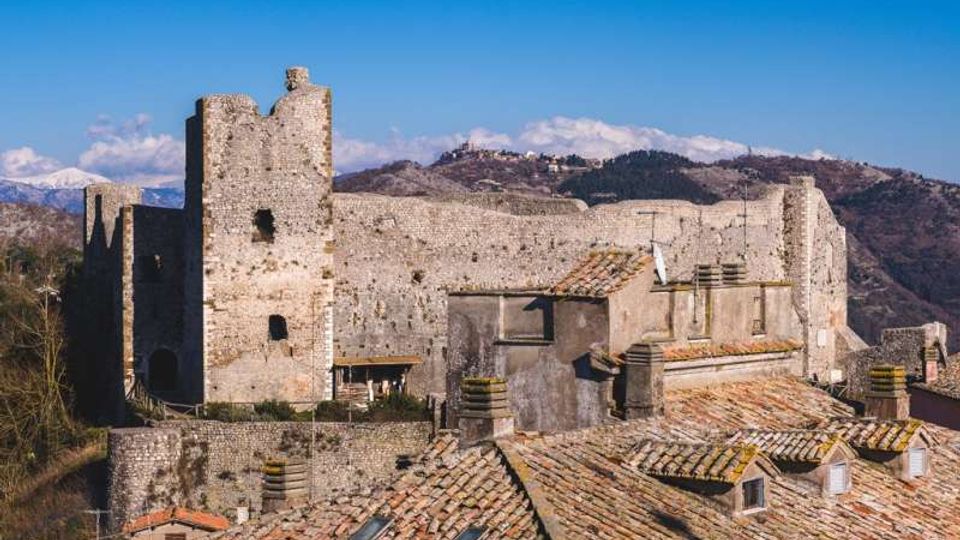 Castello medievale in pietra su un’altura, con alte mura e una torre quadrata. In primo piano si vedono tetti di case con coppi in terracotta, sullo sfondo colline e montagne sotto cielo sereno.