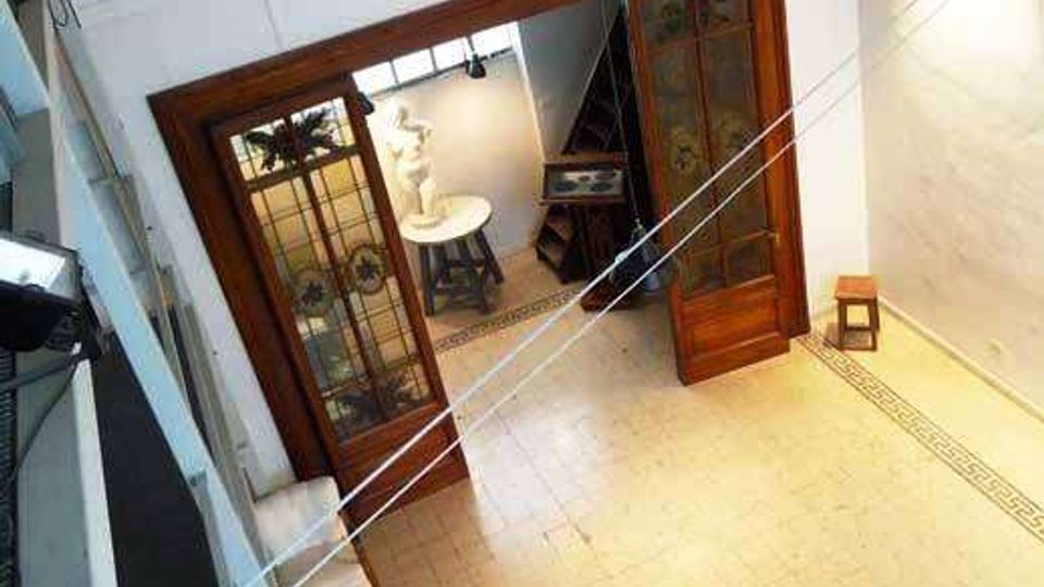 Interno di una galleria o museo visto dall’alto, con pavimento chiaro a piastrelle e grandi porte in legno con pannelli vetrati e decorati che conducono a una sala illuminata. Sulla parete superiore sono allineati busti decorativi e l’ambiente è illuminato da faretti, con un tavolo e piccoli arredi lungo il perimetro.