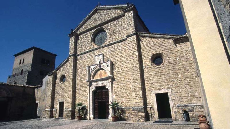 Facciata in pietra di una chiesa storica con frontone triangolare, rosone circolare e portale decorato con architrave scolpito. Davanti si apre una piazza lastricata; sullo sfondo a sinistra si vede una torre in pietra sotto un cielo sereno.