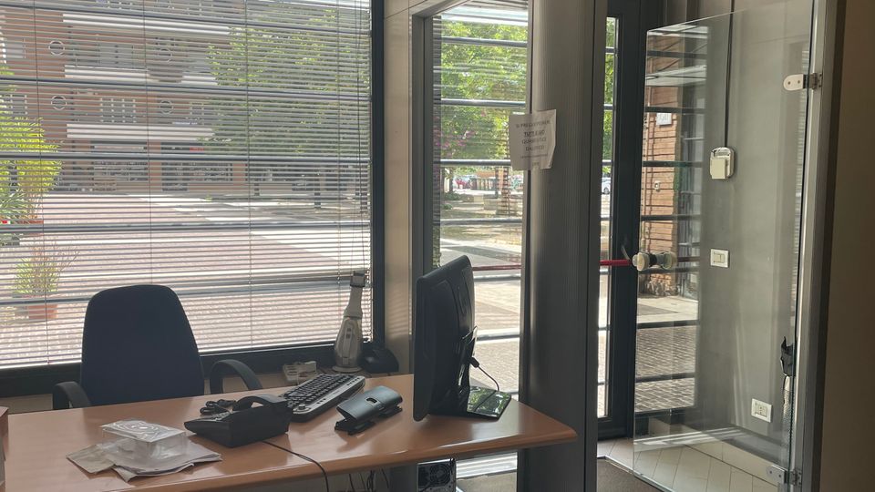Interno di un piccolo ufficio con scrivania, sedie e monitor, affacciato su grandi finestre con veneziane. Accanto c’è un ingresso vetrato con telaio scuro e una colonna centrale, con vista sulla strada esterna.
