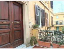 Ingresso di un edificio residenziale con grande portone in legno scuro e facciata color crema, affiancato da finestre con persiane scure e fioriere. In primo piano un piccolo balcone o pianerottolo con ringhiera metallica e vasi rettangolari di piante, con altri palazzi sullo sfondo.