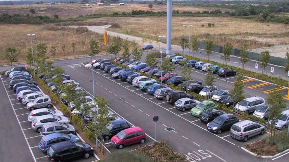 Ampio parcheggio asfaltato con file di auto negli stalli, corsie centrali e segnaletica orizzontale “STOP”, con aiuole e giovani alberi lungo i bordi. Sullo sfondo si vedono campi aperti e una torre con insegna pubblicitaria vicino alla strada d’accesso.