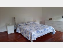 Camera da letto luminosa con pareti bianche e pavimento in legno scuro, con letto matrimoniale al centro coperto da copriletto bianco a fiori blu. Ai lati ci sono comodini bianchi e sulla parete a destra una cassettiera bianca lunga con una cornice appoggiata sopra.