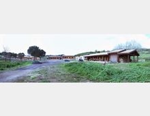 Ampio piazzale sterrato in una zona rurale, circondato da prati e leggere colline, con diversi edifici bassi e lunghi in legno e muratura. In primo piano si vede una strada in ghiaia e, sullo sfondo, tettoie aperte con cataste di legna e qualche veicolo parcheggiato.