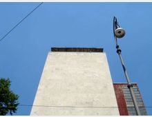 Vista dal basso di una torre rettangolare in muratura chiara con sommità sporgente, contro un cielo azzurro. A destra si vede un lampione alto e, sullo sfondo, una facciata rossa con finestre verticali.